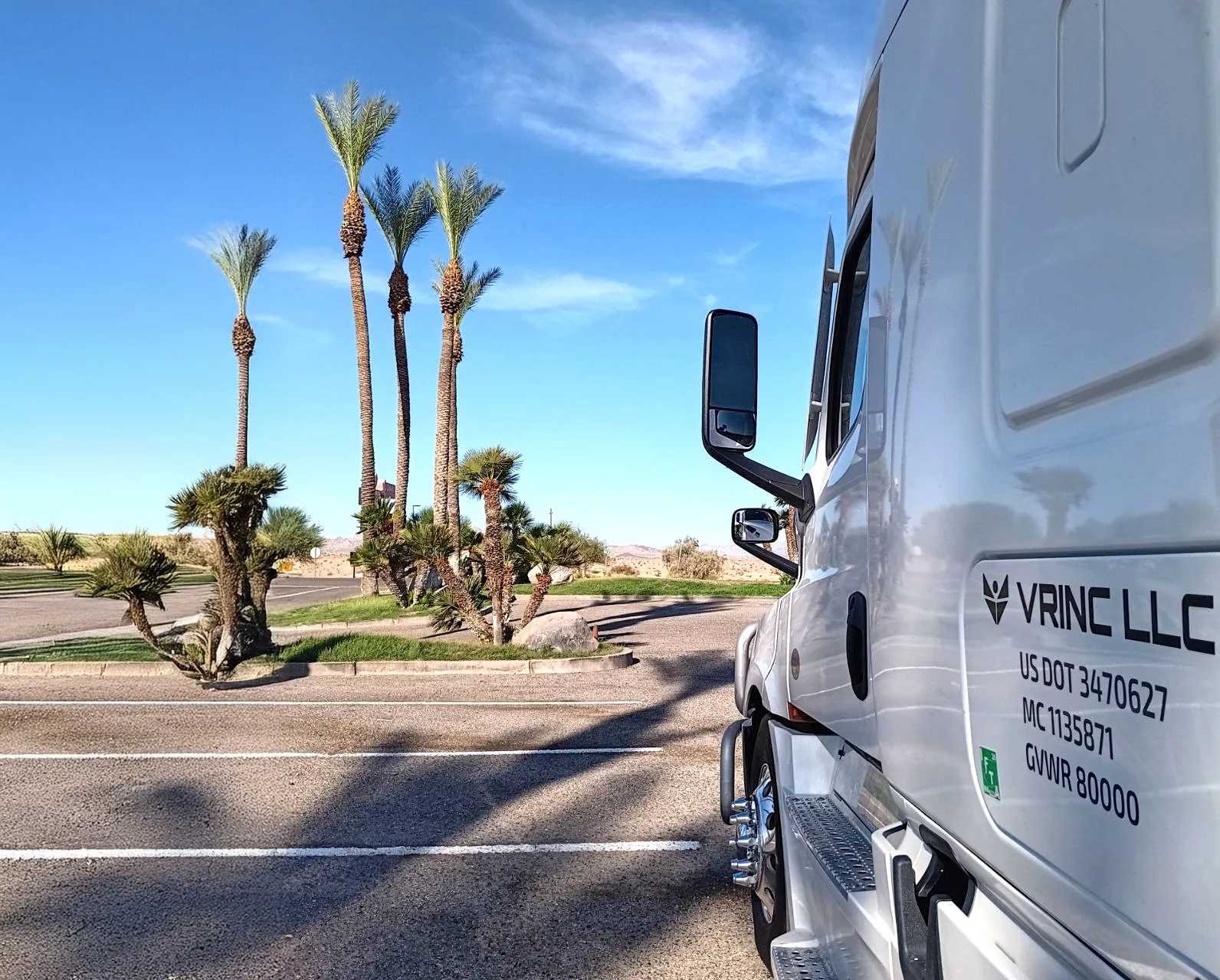 A white Vrinc LLC tractor with the DOT and MC numbers on the door, parked by palm trees under a blue desert sky.