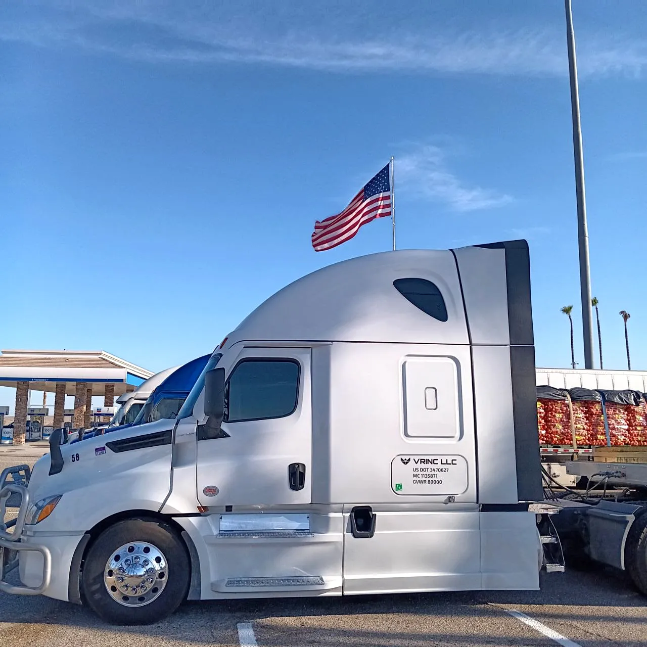 A white Vrinc LLC sleeper coupled to a tarped flatbed load, parked under an American flag at a truck stop.