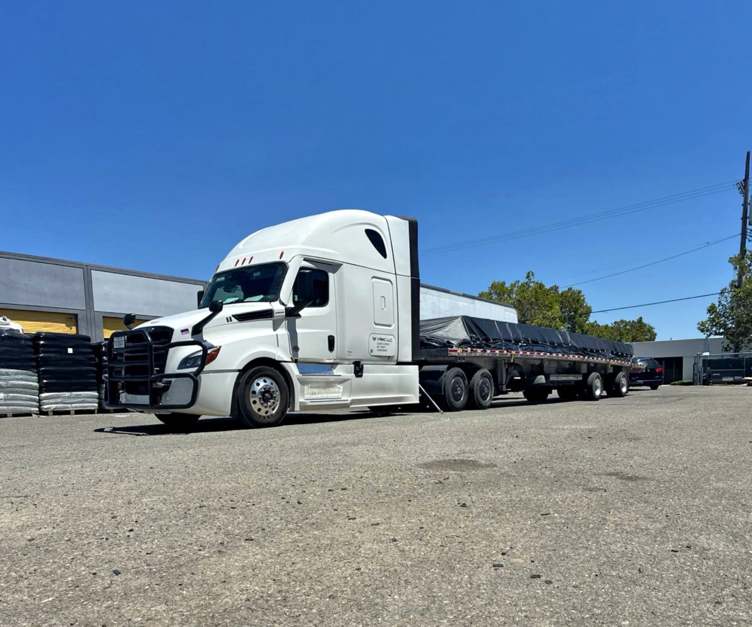A white Vrinc tractor coupled to a loaded flatbed, parked in a yard under a clear sky.