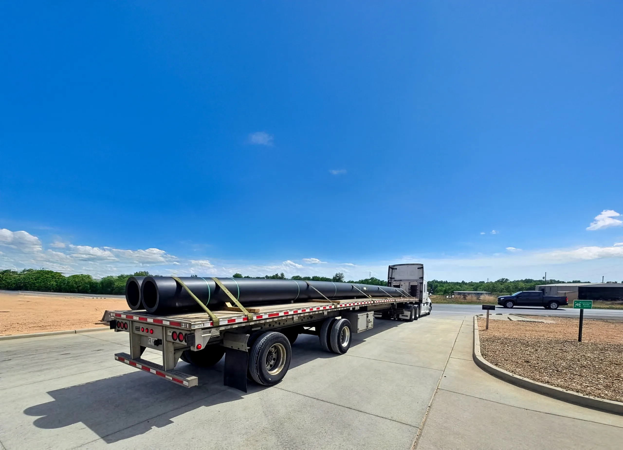 A flatbed carrying two long black pipes under straps, tractor at the far end, on a concrete lot under open sky.