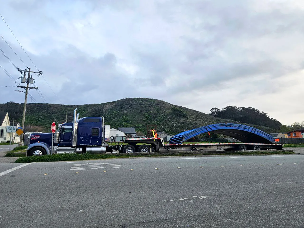 A dark blue Kenworth with a step deck trailer carrying a long curved blue steel arch, stopped on a road below a green hill.