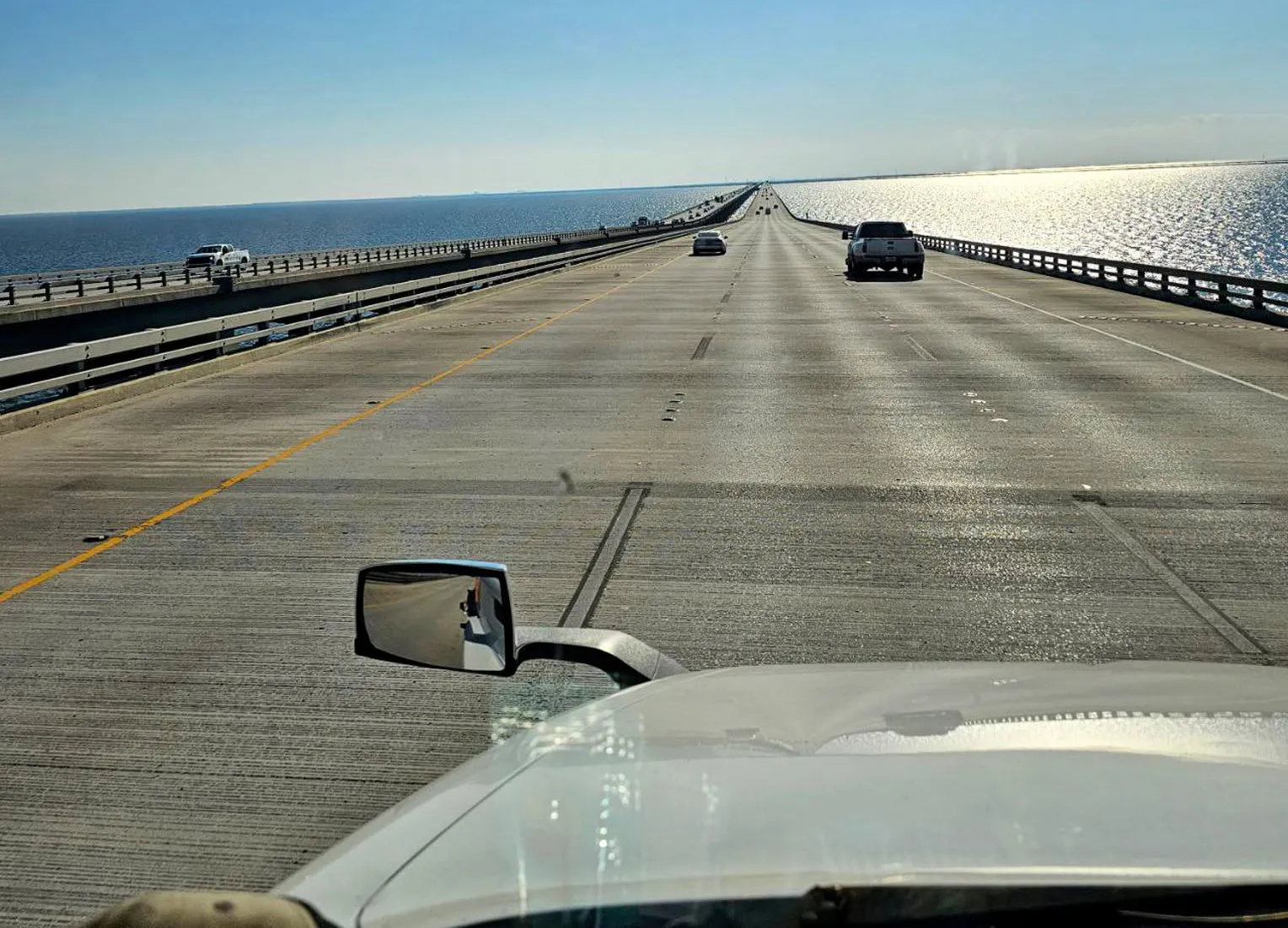 A driver's view over the white hood of a truck crossing a long bridge above open water under a clear sky.